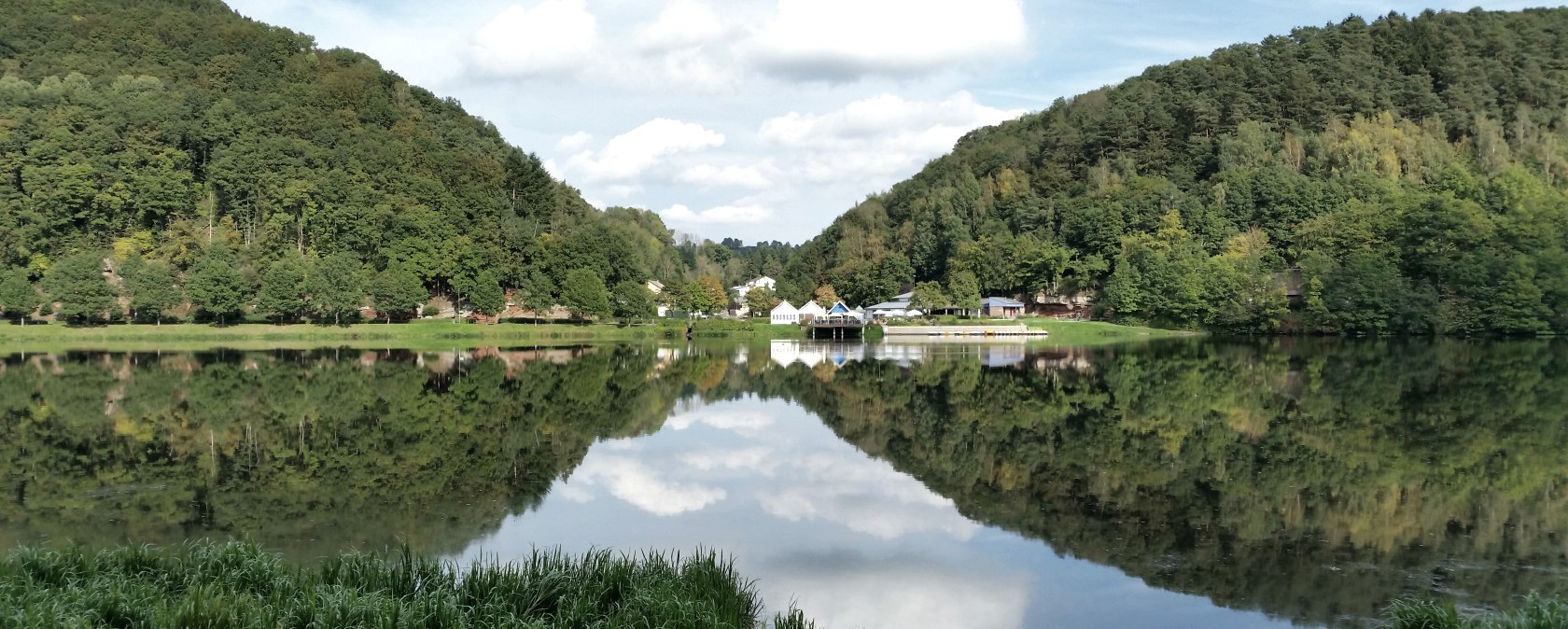 Panorama des Stausee Bitburg mit bewaldeten H&uuml;geln, Geb&auml;uden am Ufer und klarer Spiegelung im Wasser unter blauem Himmel., &copy; TI Bitburger Land