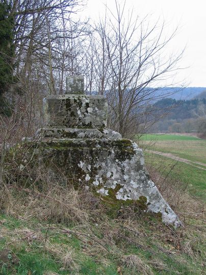 An abandoned, moss-covered stone amidst tall grass and sparsely growing trees. In the background, a green meadow and gentle hills stretch out.