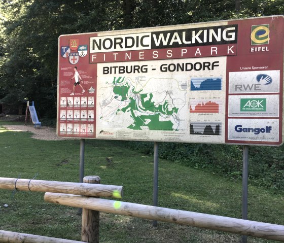 Schild des Nordic Walking Fitnessparks Bitburg-Gondorf mit Karte, Streckenprofilen und Sponsorenlogos. Im Hintergrund ein Spielplatz mit Rutsche., &copy; TI Bitburger Land