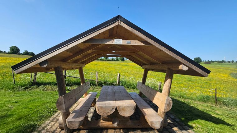 A wooden shelter with a large table and benches stands on a meadow. In the background, green fields and a clear blue sky are visible.