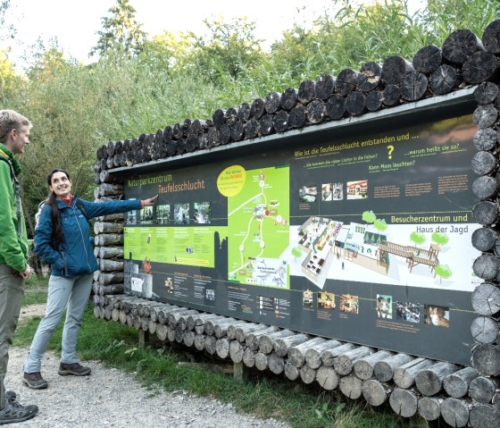 Twee mensen staan voor een informatiebord in het natuurparkcentrum Teufelsschlucht. Hierop staan kaarten en informatie over de omgeving.