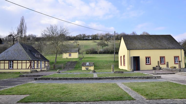 A tranquil landscape with several buildings, including a traditional house and a modern house. In the background, gentle hills and a green meadow can be seen.