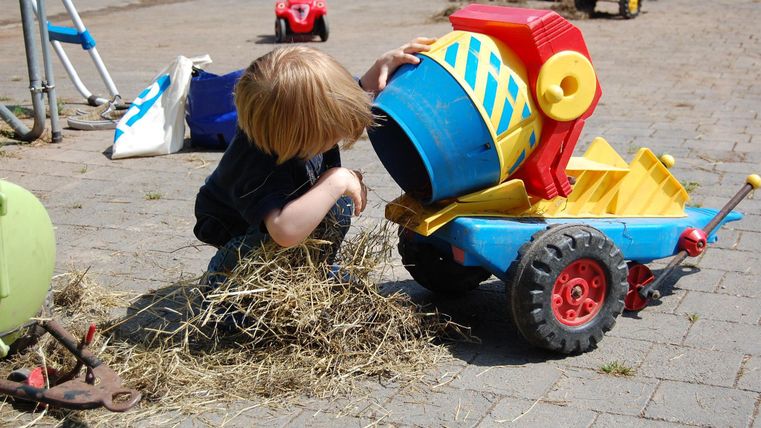A small child is playing with a colorful toy truck and a shovel. They seem to be exploring hay or grass and have tipped the bucket forward.
