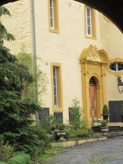 Ein historisches Gebäude mit einer gelben Fassade und einer dekorativen Eingangstür. Umgeben von Grün und Pflanzen bietet es einen einladenden Eindruck.