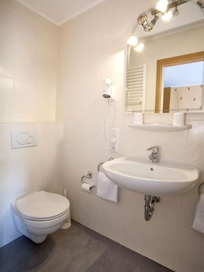 A modern bathroom with a toilet, a sink, and a large mirror. The walls are tiled in neutral colors and the floor has a gray surface.