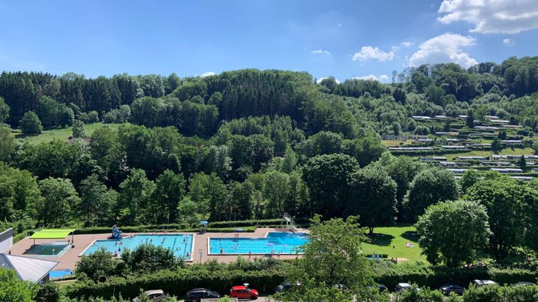 A beautiful view of a swimming pool surrounded by lush greenery and trees. The sky is blue with a few clouds.