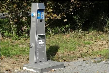 Ein Parkautomaten in einer grünen Umgebung. Er steht auf einem Schotterweg neben einer Rasenfläche.