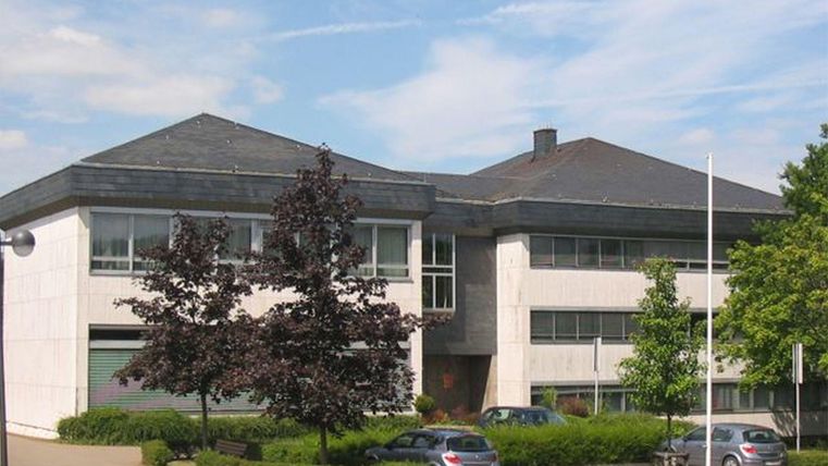 Un bâtiment moderne avec un toit à deux versants et de grandes fenêtres. Entouré d'arbres et de quelques voitures garées.