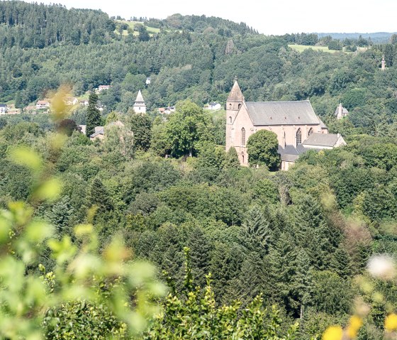 Blick auf eine Kirche, umgeben von dichten W&auml;ldern und H&uuml;geln, unter blauem Himmel. Im Vordergrund sind unscharfe Pflanzen zu sehen., &copy; Tourist-Info Bitburger Land M.Mayer