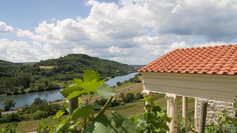 Un paysage pittoresque avec une rivière et des collines douces. Au premier plan, on voit un bâtiment avec un toit en tuiles rouges et des vignes.