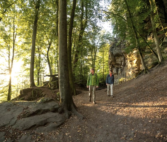 Deux randonneurs marchent sur un chemin forestier le long d'une paroi rocheuse. Le soleil brille &agrave; travers les arbres et baigne la sc&egrave;ne d'une lumi&egrave;re chaude., &copy; Dominik Ketz