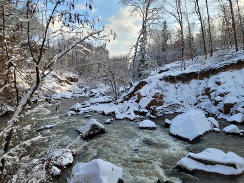 Ein malerischer Fluss fließt durch eine winterliche Landschaft mit schneebedeckten Bäumen und Felsen. Die ruhige Szene strahlt eine friedliche Atmosphäre aus.