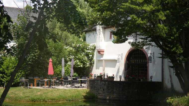 Een gezellig restaurant met een terras in de buitenlucht, omgeven door groen. Rode parasols staan klaar terwijl bomen op de achtergrond schaduw bieden.