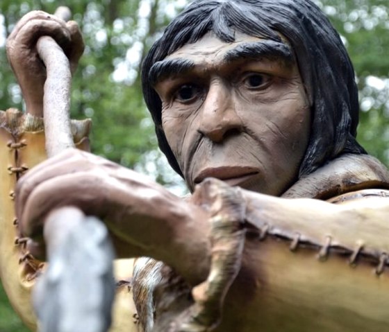 Personnage pr&eacute;historique avec une lance dans le Dinopark, entour&eacute; d'arbres. Interface Audiotour visible.