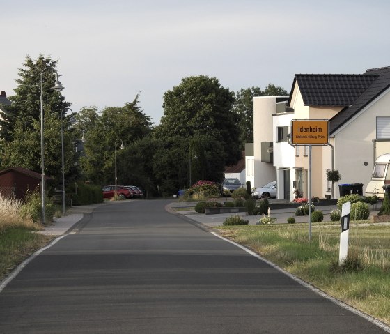 Ortseingang von Idenheim mit einem gelben Ortsschild, modernen Wohnh&auml;usern und einer ruhigen Stra&szlig;e, umgeben von B&auml;umen und Gr&uuml;nfl&auml;chen., &copy; Thomas Neises