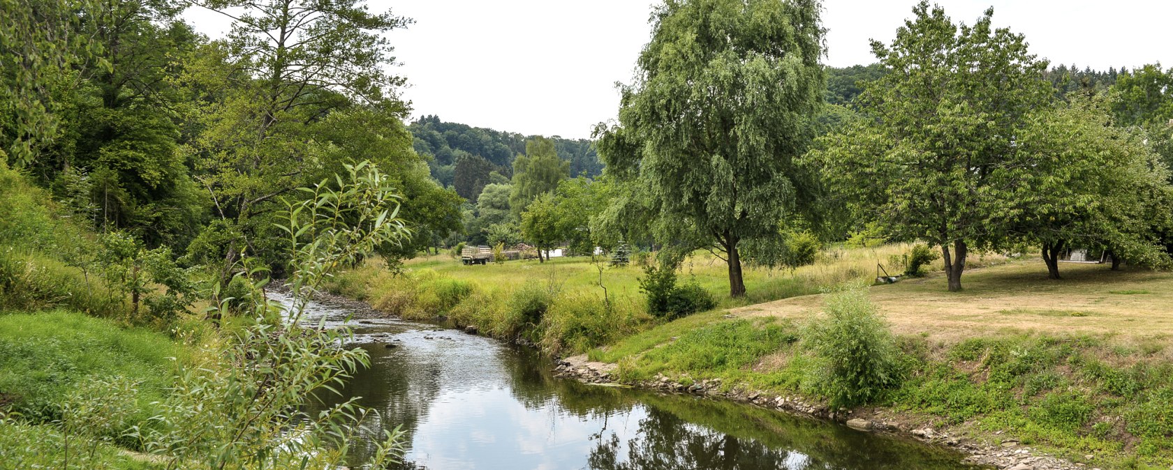 Een kleine rivier kronkelt door een groen landschap van bomen en weiden. De lucht is licht bewolkt., &copy; TI Bitburger Land