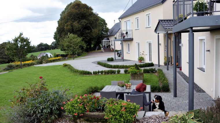 A quiet residential area with a modern house and a well-kept garden. In the foreground, there is a seating area and a dog.