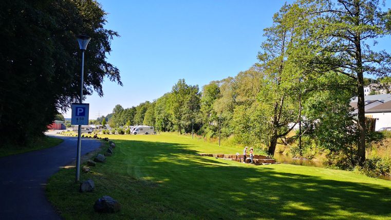 A quiet path along a green area with trees and a clear sky. On the left side, there is a parking lot.