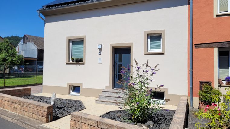 A modern house with a bright facade and a small front garden. The entrance is accessible by steps and there are some plants in front of the house.