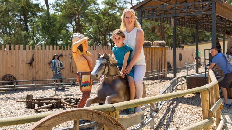 Ein Kind reitet auf einem Spielzeugpferd, während eine Frau lächelt und danebensteht. Im Hintergrund sind weitere Personen und ein Spielplatz zu sehen.