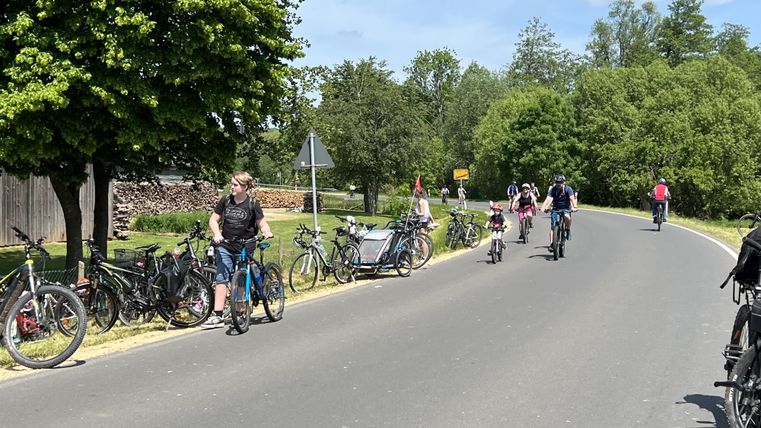 Een straat met fietsers en geparkeerde fietsen. De omgeving is groen en zonnig.
