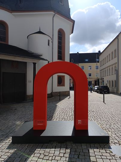 Eine große, rote Skulptur in Form eines Buchstabens "n" steht auf einem Platz. Im Hintergrund sind historische Gebäude und ein klarer Himmel zu sehen.