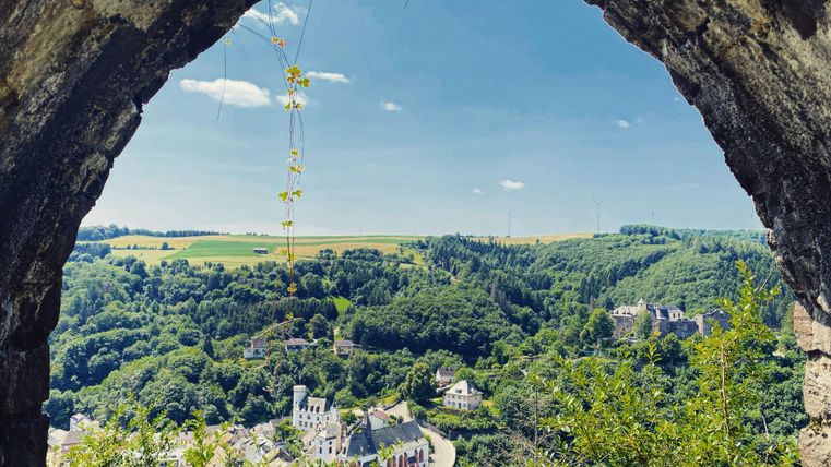 Een blik door een oude boog op een groen landschap. Op de achtergrond zijn heuvels en kleine gebouwen zichtbaar, onder een heldere blauwe lucht.