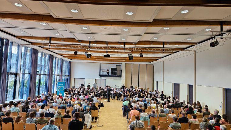 A large room with many seats is filled with an audience. On stage, an orchestra is performing in front of a large crowd.