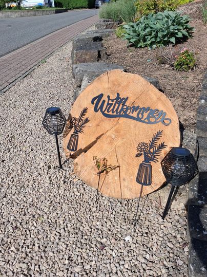 Une plaque en bois décorative avec l'inscription "Bienvenue" est posée sur du gravier. À côté de la plaque se trouvent deux lanternes noires et quelques plantes vertes.