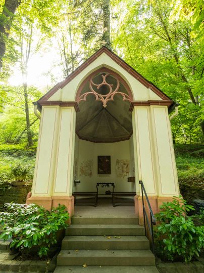 A small chapel stands in the forest, surrounded by tall trees and green foliage. The entrance has a staircase and is colorfully designed.