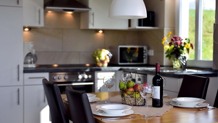 A modern kitchen with a dining table on which wine and fruit are placed. In the background, kitchen appliances and fresh flowers can be seen.