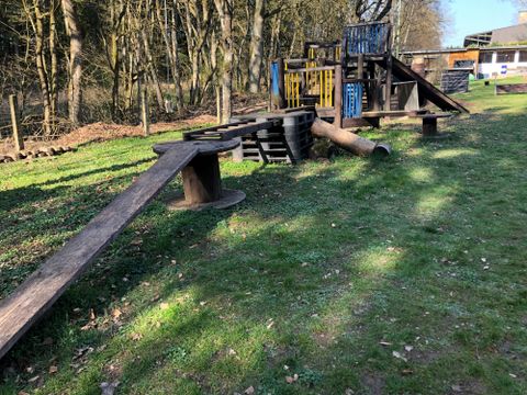 A playground with wooden structures and various climbing elements. The surroundings are green and surrounded by trees.
