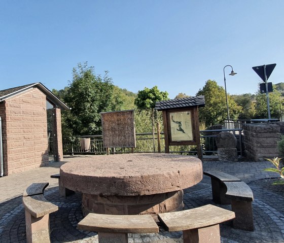 Dorpsplein in Brecht met ronde bank, informatieborden en klein gebouw. Omringd door bomen en een weg op de achtergrond., &copy; TI Bitburger Land, Steffi Wagner