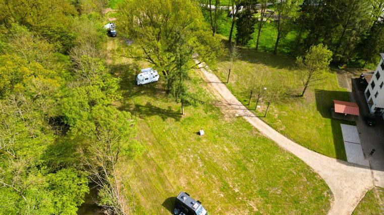Een groene weide met bomen en een parkeerplaats voor campers. Op de achtergrond loopt een pad en een gebouw is zichtbaar.