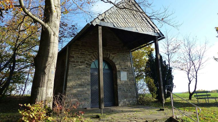 Une petite chapelle en pierre entourée d'arbres. Le ciel est clair et bleu, et en arrière-plan, il y a un banc.