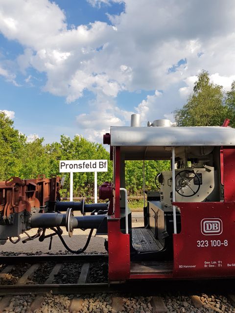 A red train is standing on the tracks at the Pronsfeld Bf station. The sky is partly cloudy with some nice clouds.
