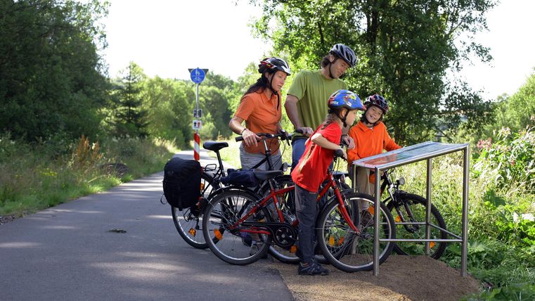 Eine Familie mit Fahrrädern steht an einem Informationsschild auf einem Radweg. Sie tragen Helme und orangefarbene Shirts und schauen auf die Umgebung.
