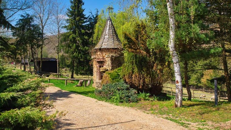 Ein idyllischer Weg führt zu einem kleinen, steinernen Gebäude mit einem spitzen Dach. Umgeben von Bäumen und Grünflächen strahlt die Szenerie Ruhe und Naturverbundenheit aus.