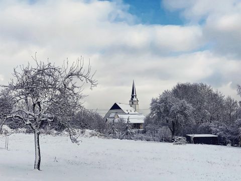 Eine winterliche Landschaft mit schneebedeckten Bäumen und einem Dorf im Hintergrund. Der Kirchturm ragt hoch in den Himmel unter einem bewölkten, blauen Himmel.