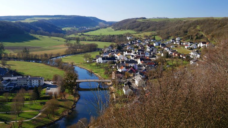 Un paysage pittoresque avec un petit village au bord de la rivière. Des prairies vertes et des collines douces entourent la scène.