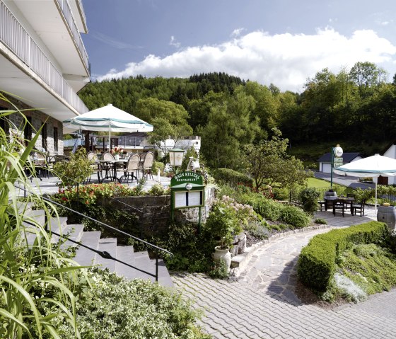 Terrasse de l'h&ocirc;tel Haus Kylltal avec tables et parasols, entour&eacute;e d'une v&eacute;g&eacute;tation luxuriante et d'une for&ecirc;t en arri&egrave;re-plan., &copy; Hotel Haus Kylltal