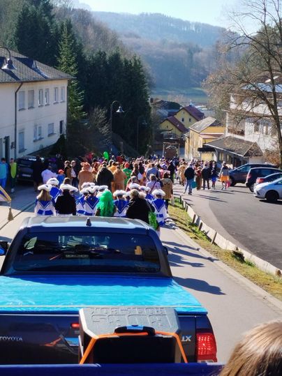 A colorful crowd is celebrating an event on a street. In the background, houses and nature are visible.