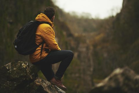 A man is sitting on a rock and looking into a green gorge. He is wearing a yellow jacket and has a backpack with him.