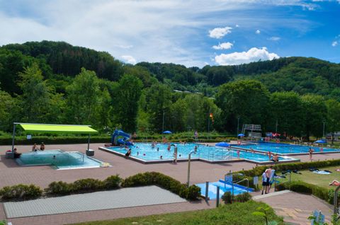 A swimming pool with multiple basins and many people relaxing and swimming. The surroundings are green and offer beautiful landscapes under a blue sky.