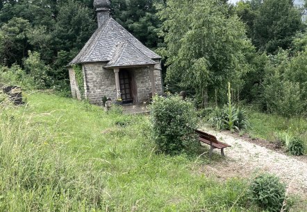 Une petite chapelle en pierre avec un toit en ardoise se dresse au milieu d'une v&eacute;g&eacute;tation luxuriante. Un chemin de gravier m&egrave;ne &agrave; un banc en bois devant la chapelle., &copy; Daniel K&ouml;hler