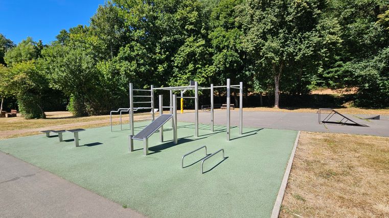 Ein Trainingsbereich im Freien mit verschiedenen Fitnessgeräten. Umgeben von Bäumen und Rasen entsteht eine einladende Atmosphäre.