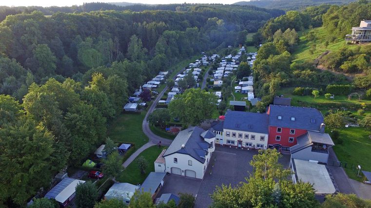 Een schilderachtige camping omgeven door groene bomen. Op de achtergrond zijn talloze campers en tenten te zien.