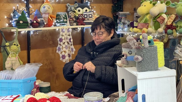 Een vrouw zit aan een tafel en breit. Op de achtergrond zijn kleurrijke handwerken en speelgoed te zien.