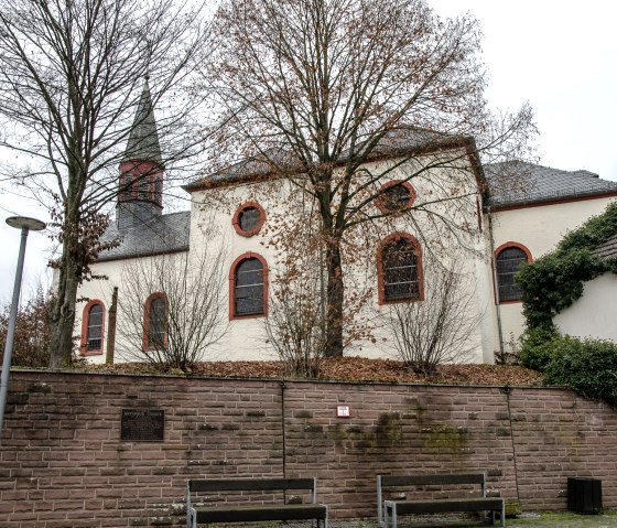 Eine Kirche in Wi&szlig;mannsdorf mit roten Fensterrahmen, umgeben von kahlen B&auml;umen und einer Steinmauer im Vordergrund., &copy; Tourist-Info Bitburger Land