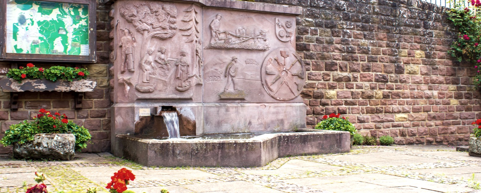 Une fontaine de village en pierre avec des reliefs et de l'eau qui coule, entour&eacute;e de fleurs rouges et d'un panneau d'information sur un mur en pierre., &copy; TI Bitburger Land - M. Mayer